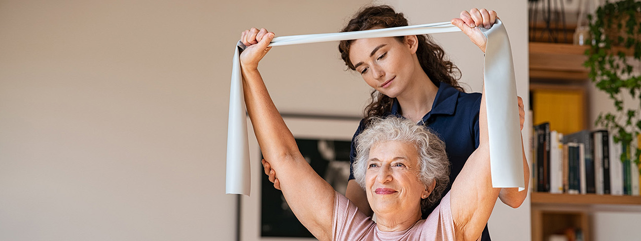 Young trainer helping senior woman with resistance band. Lovely physiotherapy health worker exercise with old woman at home. Happy smiling senior woman using rubber band with coach to improve her muscle tone.